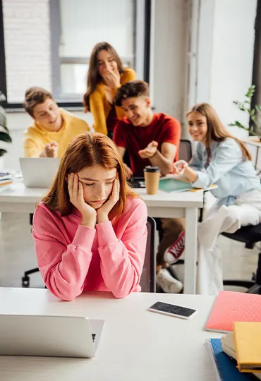 Imagen que muestra delante a una chica en un aula expresando tristeza y detrás a un grupo de chicos y chicas señalándola y riéndose.